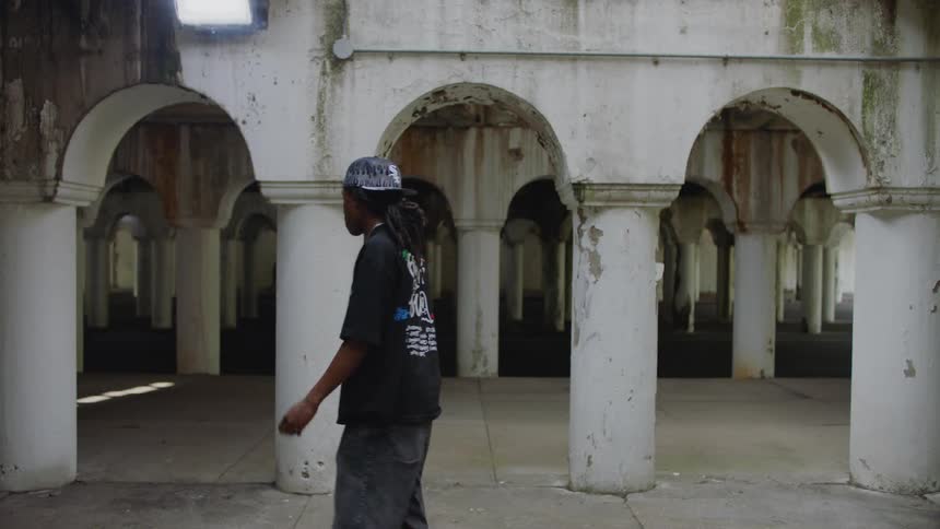 A young man with dreadlocks dances in slow motion in an abandoned, dilapidated building with white pillars and arches