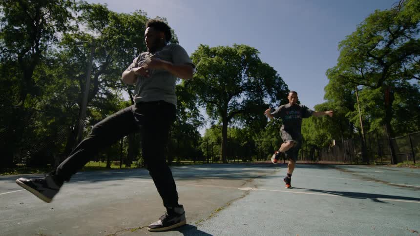 Dynamic slow-motion footage of two male dancers performing a complex footwork routine on a sunlit park court
