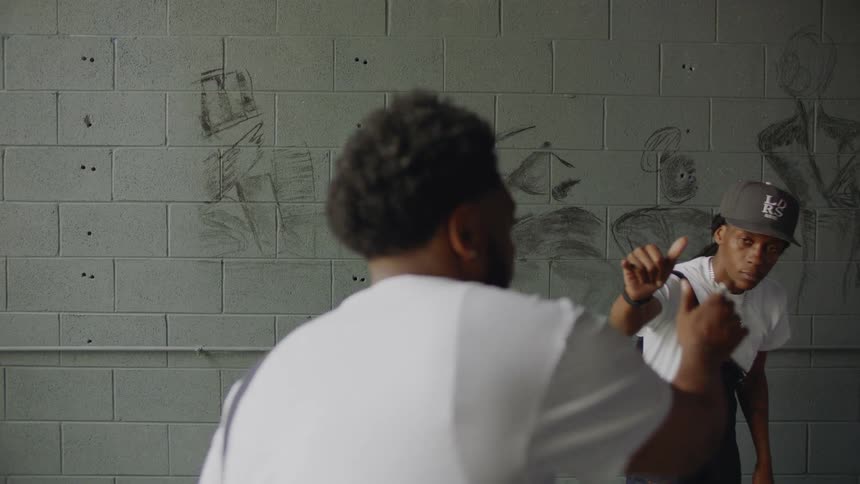 A young Black man with a beard raps to the camera and points to his grillz while his friend stands in the background