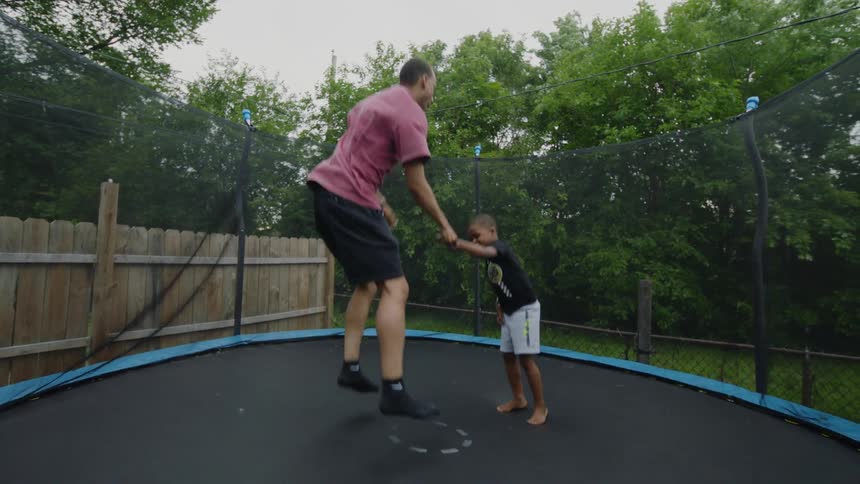 A father and his young son have fun jumping together on a trampoline in their backyard in slow motion