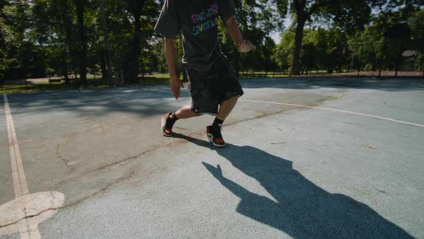 A man performs intricate footwork and dance moves in slow motion on an outdoor basketball court in a sunny park