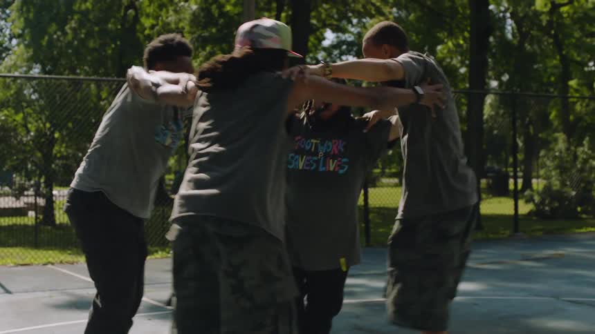Four male dancers perform an energetic and synchronized Chicago footwork dance routine on an outdoor basketball court