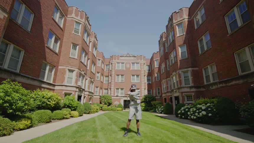 A young man dances with energy and style in the grassy courtyard of a classic brick apartment complex on a sunny day