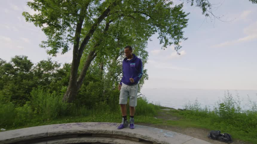 A stylish man performs an expressive, fluid dance in slow motion on a circular stone structure in a park overlooking the water
