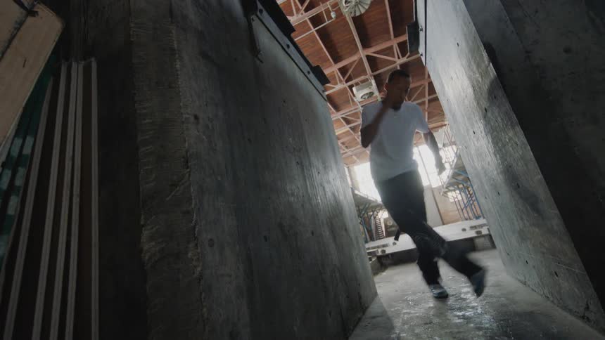 A man performs energetic hip-hop and freestyle dance moves in a narrow industrial hallway between two large concrete blocks