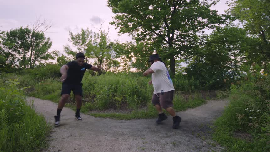 Two male friends perform synchronized hip-hop dance moves in slow motion on a forked trail in a park at dusk