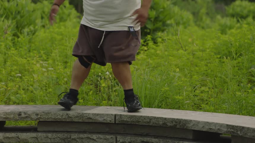 A person wearing a bucket hat dances expressively in slow motion in a lush green outdoor park setting