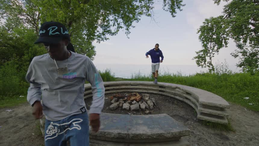 Two men dance in slow motion around a stone fire pit in a park overlooking the water