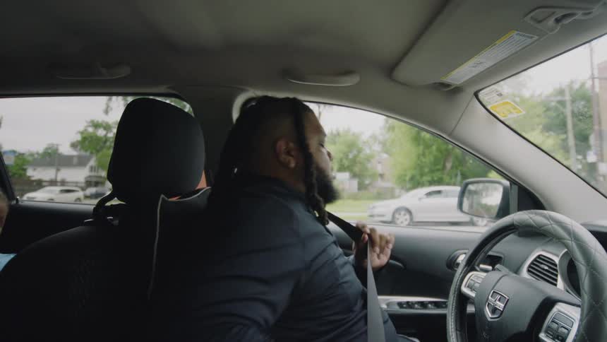 A man with dreadlocks gets into the driver's seat of his car, buckles his seatbelt, and begins to drive in slow motion