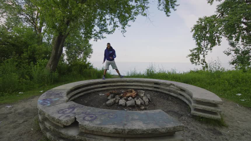 A man in a purple sweatshirt performs an energetic freestyle dance in slow motion on a graffiti-covered fire pit overlooking the water