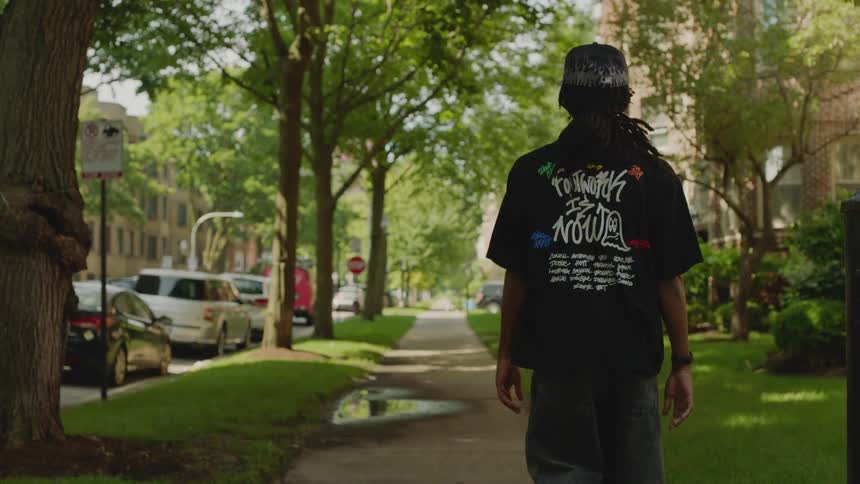 Back view of a man with long dreadlocks walking away from the camera in slow motion down a tree-lined city sidewalk