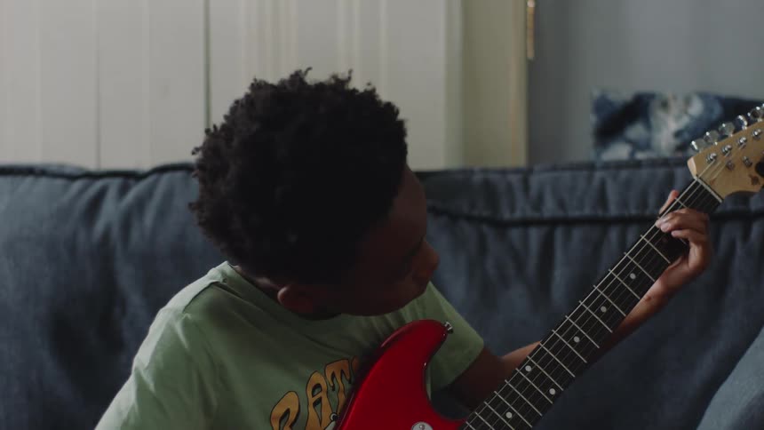 A young African American boy sits on a couch and concentrates on learning to play a red electric guitar