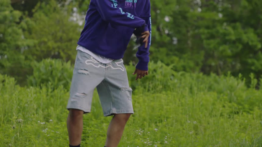 A man in a purple hoodie and sneakers performs an energetic footwork dance routine on a stone ledge in a park