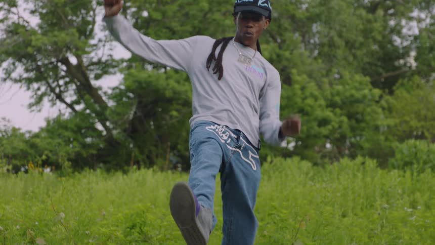 A stylish young man with dreadlocks performs an energetic hip-hop dance routine in a lush green park