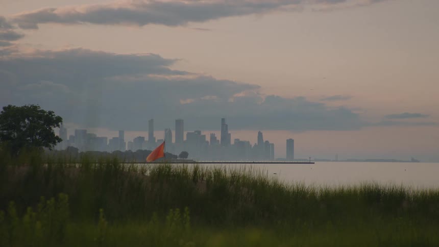 A hazy view of the Chicago skyline across Lake Michigan at sunrise, with a grassy shore and red flag in the foreground