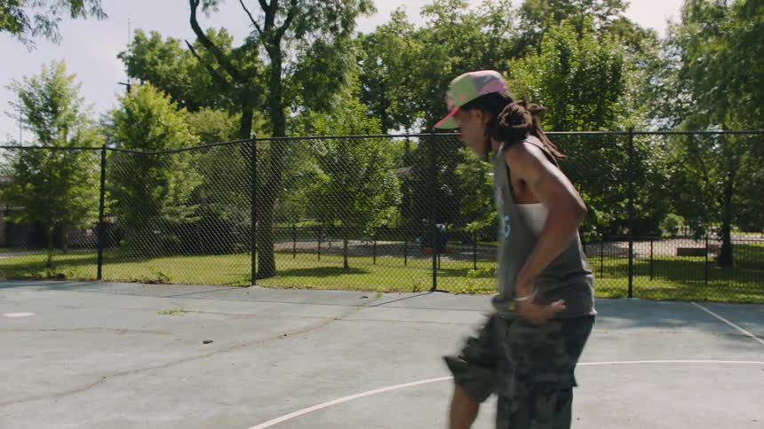 A talented dancer performs an energetic footwork routine on an outdoor basketball court as his friends watch in slow motion
