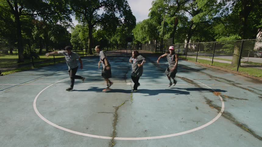 A diverse group of four male dancers perform an energetic synchronized hip-hop routine on an outdoor basketball court in a city park