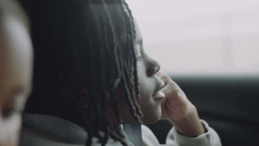 A pensive young Black child with dreadlocks gazes out the window on a contemplative car journey