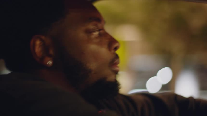 A contemplative man drives through the city at night, his face illuminated by the passing street lights and traffic