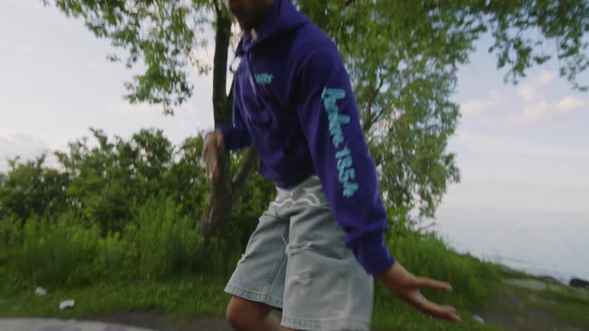 A stylish young man performs an energetic hip-hop dance routine in slow motion on a graffiti-covered stone fire pit by the lake