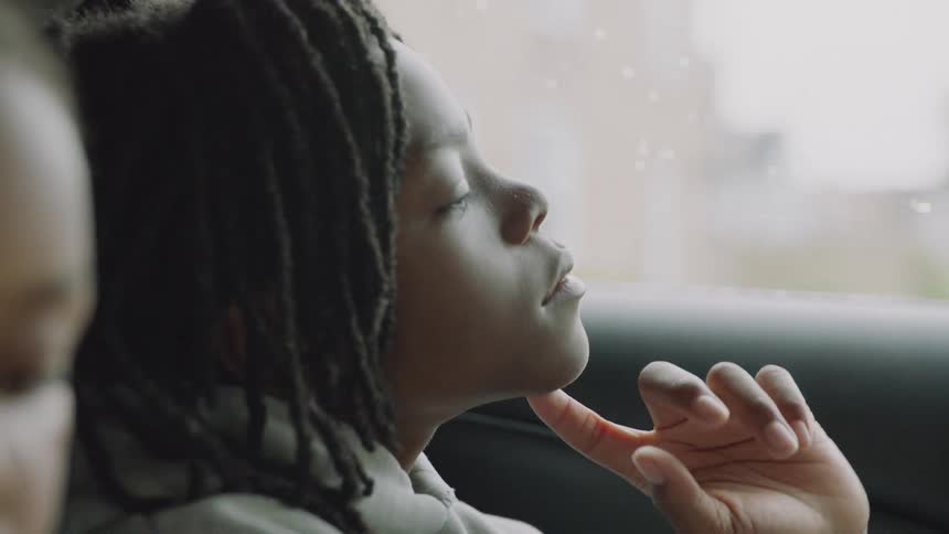 A pensive young child with dreadlocks looks out of a car window in slow motion on a rainy day