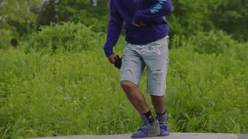 An expressive male dancer performs intricate footwork in slow motion on a stone wall in a lush green park