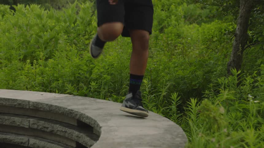 A stylish young man performs intricate footwork dance moves in slow motion on a stone ledge in a park