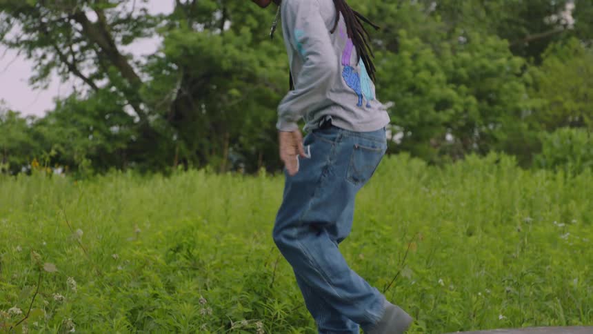 Slow motion shot of a young man with dreadlocks dancing freestyle hip-hop on a stone ledge surrounded by a lush green meadow