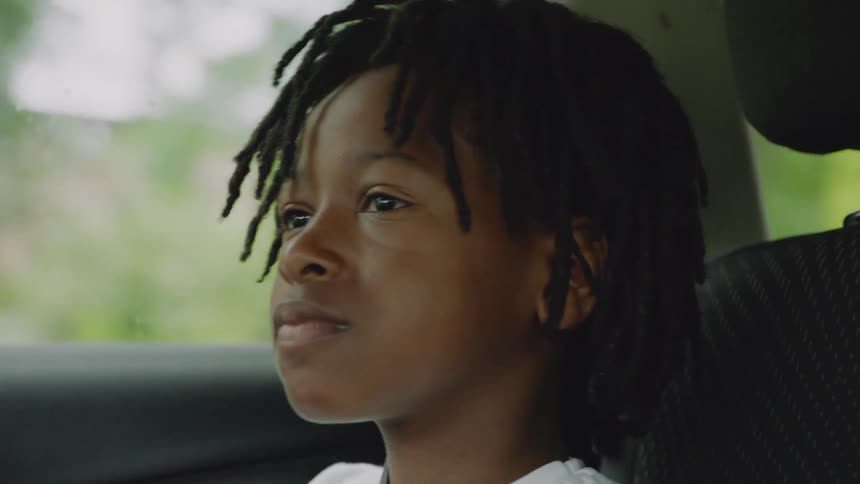 A pensive young boy with dreadlocks looks out the car window during a ride, his expression shifting from thoughtful to a happy smile