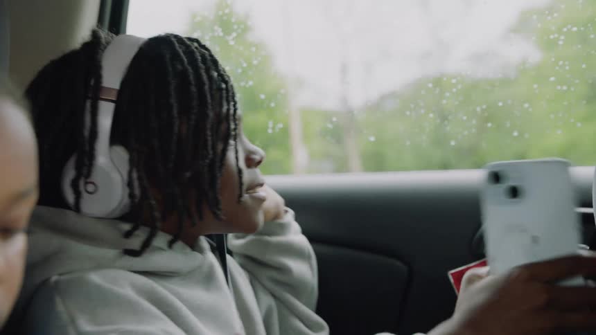 A contemplative young boy listens to music on his headphones while looking out the window during a car ride on a rainy day