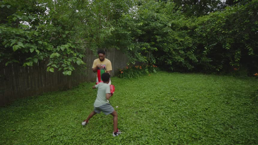 A loving father teaches his young son how to box in their lush green backyard, holding a punching shield for practice