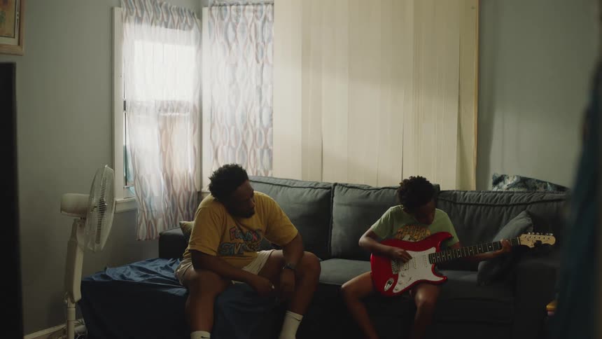 Supportive Black father watches his son practice playing the red electric guitar on the couch in their living room