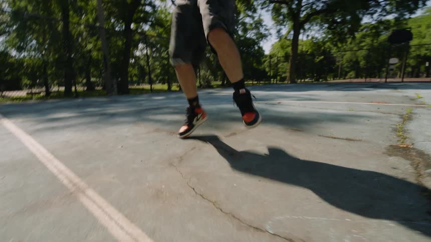 A young Black man energetically performs a dynamic footwork dance routine on an outdoor basketball court on a sunny day