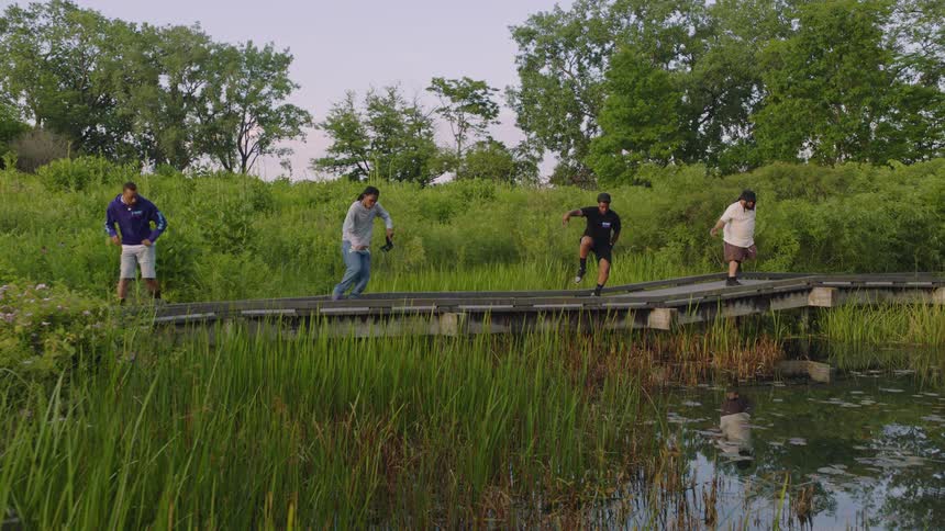 Four friends perform a synchronized dance routine in slow motion on a boardwalk over a tranquil pond in a park