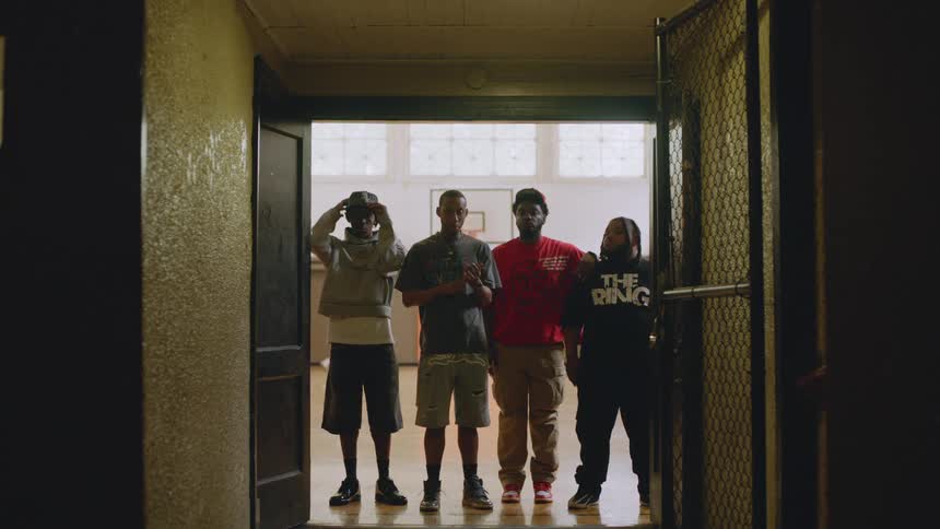 A group of four young Black men stand together in a doorway, silhouetted against the bright light of an indoor basketball court