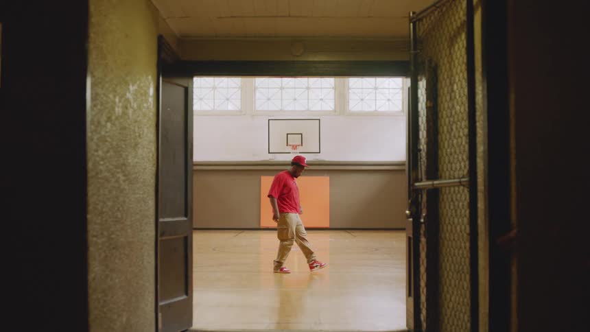 Two male hip-hop dancers practice smooth moves in slow motion on an indoor basketball court, as viewed from a dark hallway