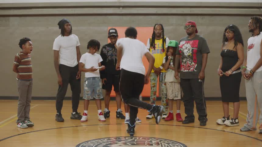 A talented young Black man performs an energetic hip-hop dance routine for a supportive crowd of adults and children in a gymnasium