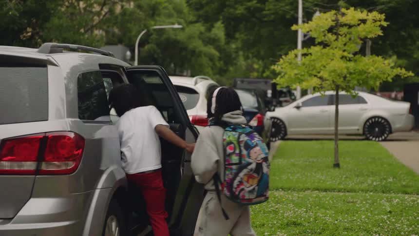 Two young African American boys with backpacks walk along a city sidewalk and get into a car after school