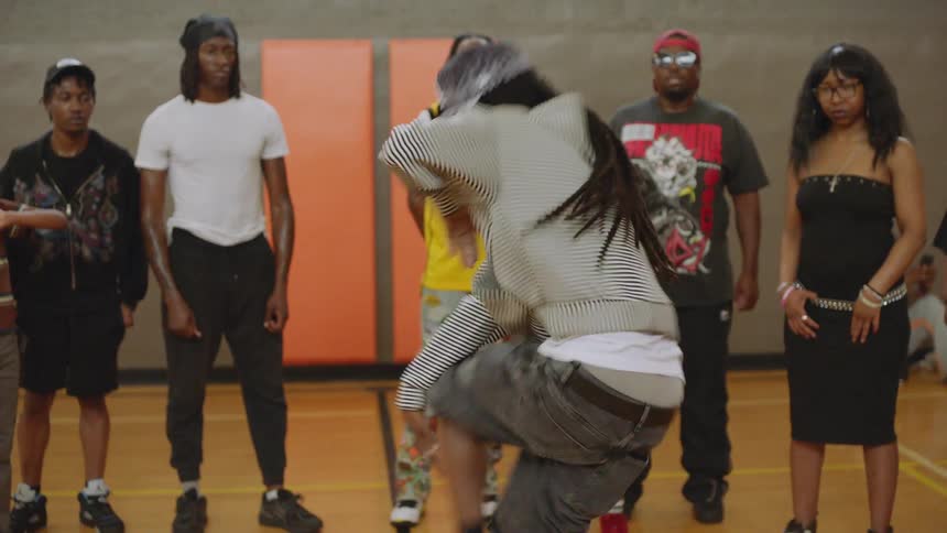 A young man with dreadlocks performs an intense and energetic street dance routine as a diverse group of people watches inside a gymnasium