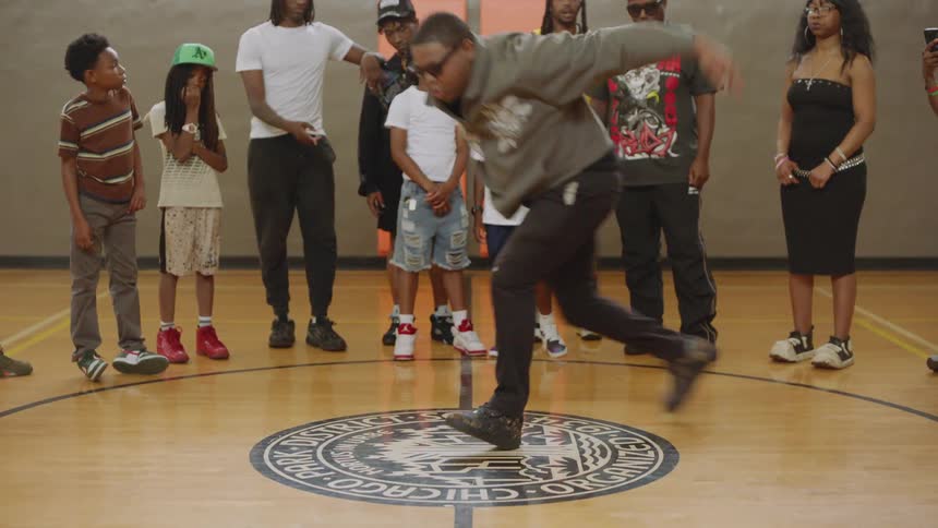 A talented man performs an incredible footwork dance routine for a crowd of adults and children on a basketball court