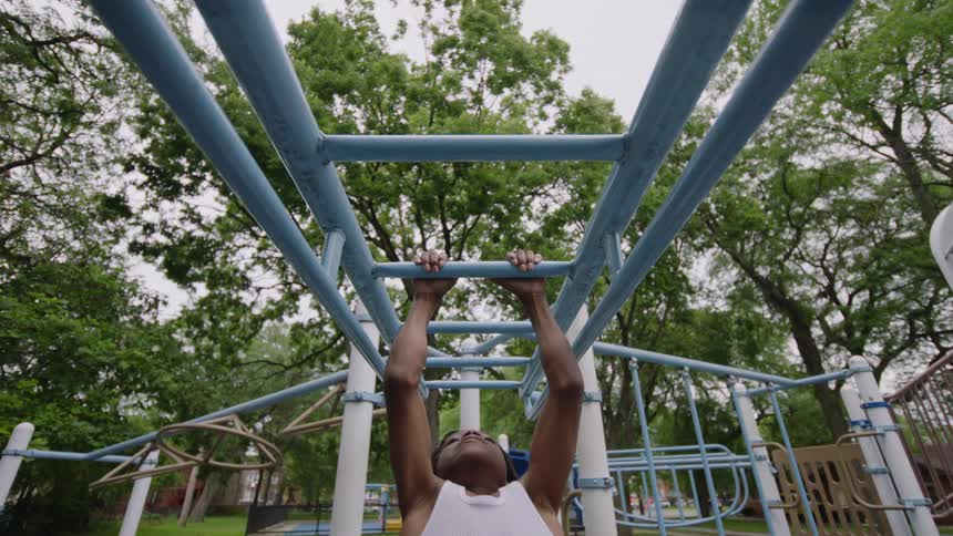 A fit young Black man performs pull-ups on the monkey bars at an outdoor park gym