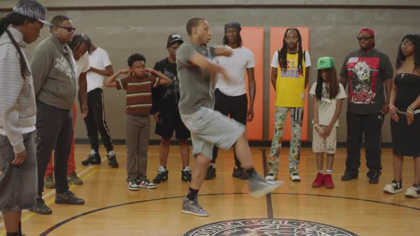 A talented young Black man performs an energetic footwork dance routine for a crowd on an indoor basketball court