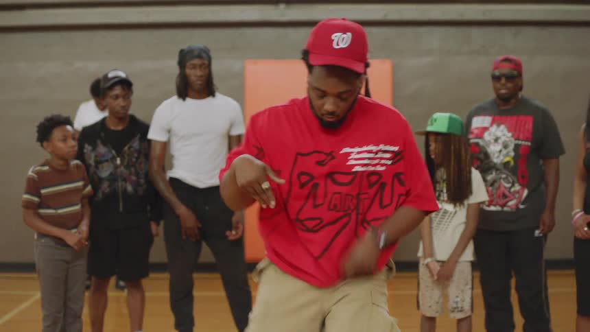 A man in a red t-shirt dances in slow motion on a basketball court while a crowd of people watches