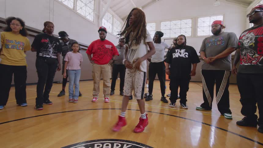 A young dancer with long dreadlocks commands the floor with dynamic street dance moves as a crowd watches in a gymnasium