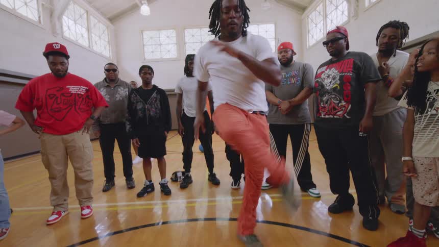 An energetic male dancer showcases powerful street dance moves in the center of a circle inside a gymnasium