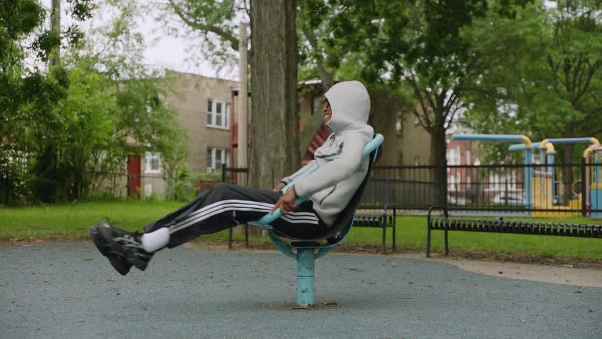 A joyful young Black man in a hoodie smiles while spinning on a playground ride in slow motion