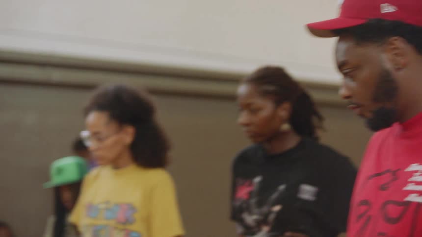 A man with long dreadlocks shows off his impressive Chicago Footwork skills as a focused crowd watches him dance in a gymnasium