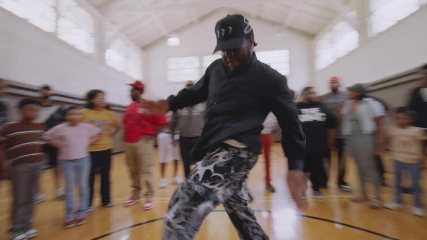 A man performs energetic Chicago Footwork dance moves in slow motion while surrounded by a crowd in a gymnasium