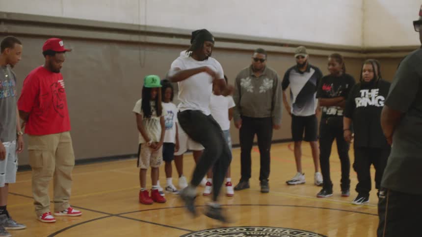 A talented young man performs an energetic footwork dance routine in a circle while a crowd watches in a community center gymnasium