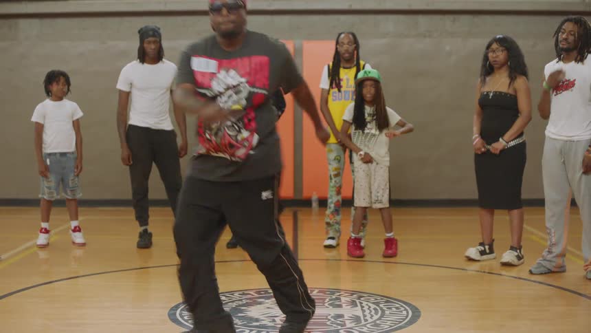 A man in sunglasses and a baseball cap performs an energetic street dance routine in the center of a basketball court while a diverse group of onlookers watch